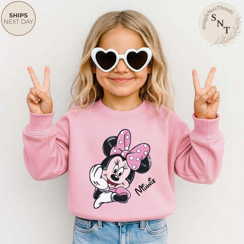 May include: A pink sweatshirt featuring a Minnie Mouse graphic with a polka dot bow. The word "Minnie" is written in cursive. The child is wearing white heart-shaped sunglasses and making a peace sign with their fingers.