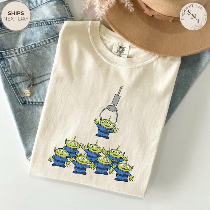 May include: Cream-colored t-shirt featuring a graphic of green aliens in blue uniforms being lifted by a claw machine. The shirt has a Comfort Colors tag. A tan hat and blue jeans are in the background.