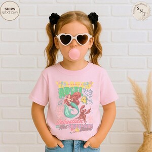 May include: A light pink short-sleeve t-shirt featuring a graphic print of Ariel, Flounder, and Sebastian. The text reads Atlantica Ocean Tour 1991, Sebastian with the Hot Crustacean Band. The design includes colorful coral and sea-themed illustrations.
