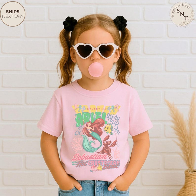 May include: A light pink short-sleeve t-shirt featuring a graphic print of Ariel, Flounder, and Sebastian. The text reads Atlantica Ocean Tour 1991, Sebastian with the Hot Crustacean Band. The design includes colorful coral and sea-themed illustrations.
