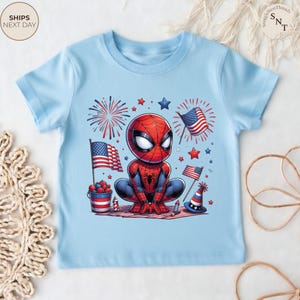 May include: A light blue short-sleeve t-shirt featuring a graphic of a cartoon Spider-Man character crouching. The design includes American flags, red and blue fireworks, stars, a bucket of berries, and a party hat in a patriotic color scheme.