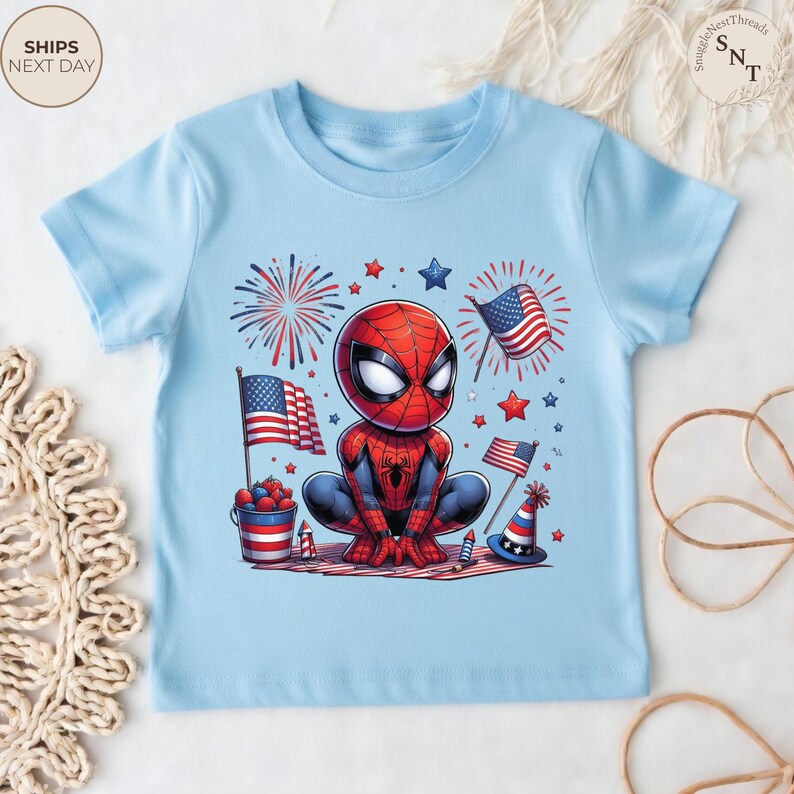 May include: A light blue short-sleeve t-shirt featuring a graphic of a cartoon Spider-Man character crouching. The design includes American flags, red and blue fireworks, stars, a bucket of berries, and a party hat in a patriotic color scheme.