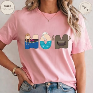 K&ouml;nnte beinhalten: Hellrosa T-Shirt mit dem Wort "MOM" im Cartoon-Stil. Das "M" zeigt eine Figur mit Z&ouml;pfen, das "O" hat ein Schneeflocken-Design und das "M" hat ein Geweih. Das Shirt hat den Text "SnuggleNestThreads" oben rechts.