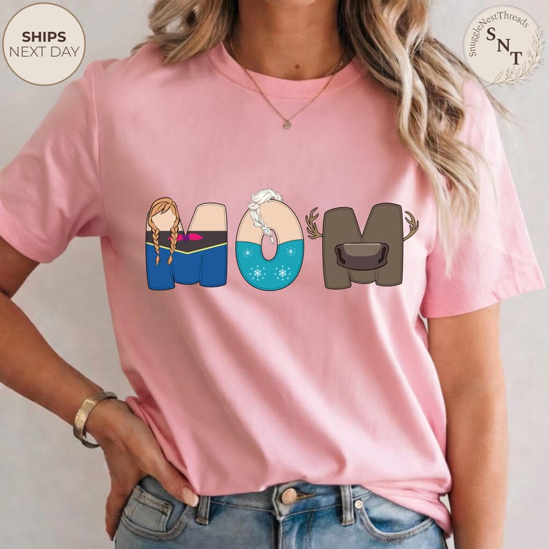 K&ouml;nnte beinhalten: Hellrosa T-Shirt mit dem Wort "MOM" im Cartoon-Stil. Das "M" zeigt eine Figur mit Z&ouml;pfen, das "O" hat ein Schneeflocken-Design und das "M" hat ein Geweih. Das Shirt hat den Text "SnuggleNestThreads" oben rechts.