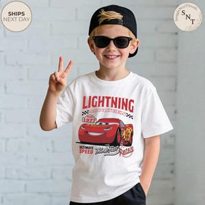 Op de afbeelding: Wit T-shirt met Lightning McQueen uit de film Cars. Het shirt heeft een rood-zwarte afbeelding van het personage met de tekst "LIGHTNING MCQUEEN" en "est. 1977". Het shirt heeft ook de zinnen "ULTIMATE SPEED" en "Think Fast, Rusteze".