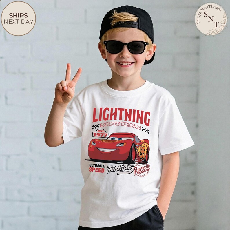 Op de afbeelding: Wit T-shirt met Lightning McQueen uit de film Cars. Het shirt heeft een rood-zwarte afbeelding van het personage met de tekst "LIGHTNING MCQUEEN" en "est. 1977". Het shirt heeft ook de zinnen "ULTIMATE SPEED" en "Think Fast, Rusteze".