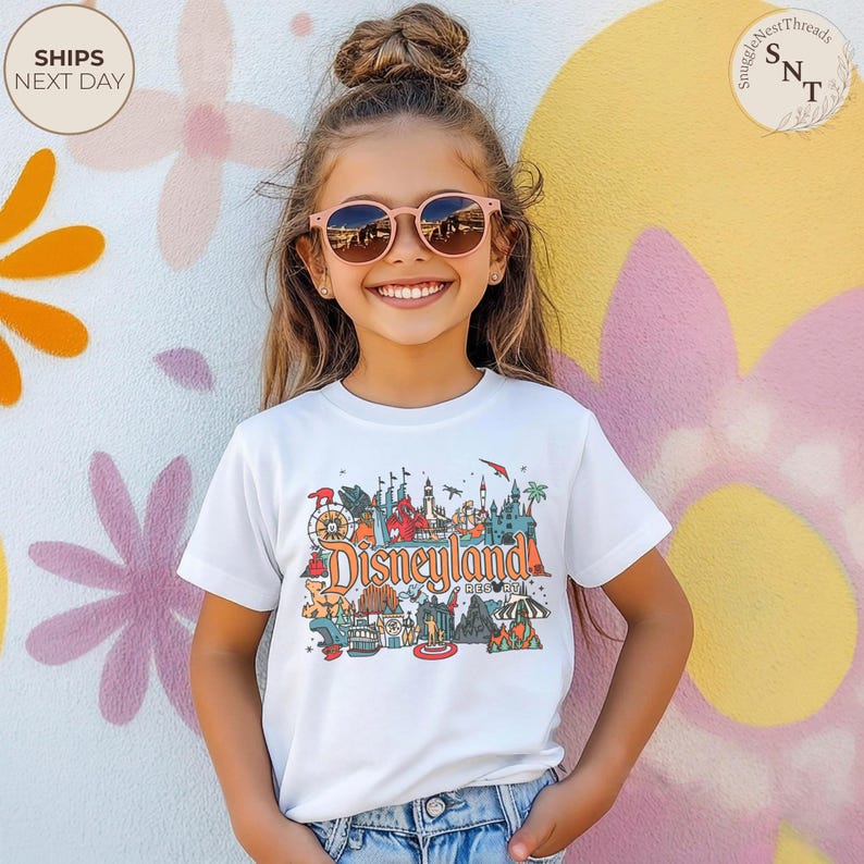 May include: A young girl wearing a white t-shirt with a colorful Disneyland graphic. The graphic features various park attractions and the text "Disneyland Resort". She is wearing pink sunglasses and smiling in front of a floral painted wall.