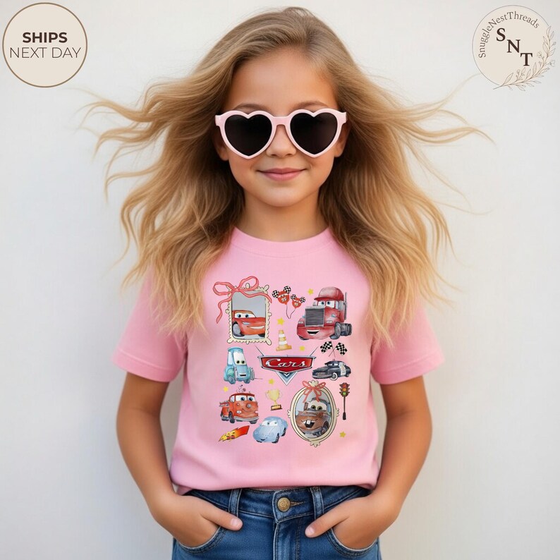 May include: A pink t-shirt featuring various Cars movie characters. The shirt has a crew neck and short sleeves. The girl is wearing heart-shaped sunglasses and blue jeans.