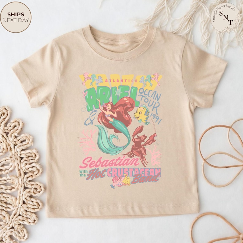 May include: A beige short-sleeve t-shirt featuring a graphic print of Ariel, Flounder, and Sebastian. The text reads Atlantica Ariel Ocean Tour 1991, Sebastian with the Hot Crustacean Band, and includes colorful illustrations of sea life and musical notes.