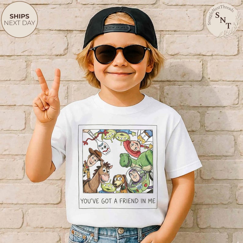 May include: White t-shirt featuring a colorful illustration of characters from the movie "Toy Story." The image includes Woody, Buzz Lightyear, and other characters. The text "YOU'VE GOT A FRIEND IN ME" is printed below the image. The child is wearing a black cap and sunglasses.
