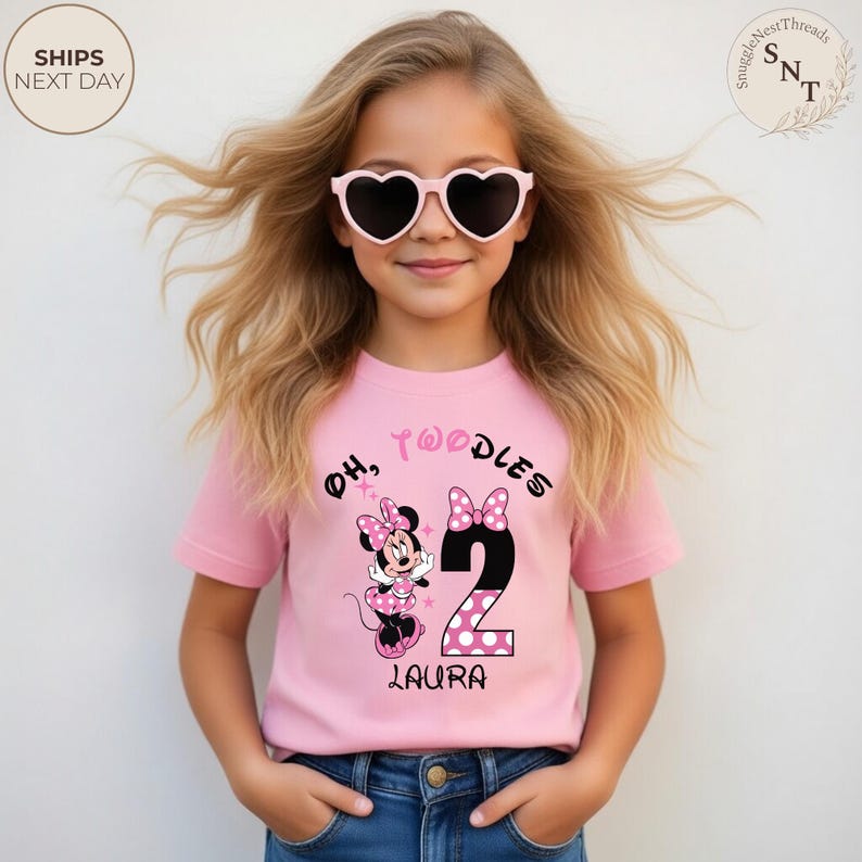 Puede incluir: Una camiseta rosa con el texto "Oh, Twoodles" y un gr&aacute;fico de Minnie Mouse. El n&uacute;mero "2" se presenta con un dise&ntilde;o de lunares. El nombre "Laura" est&aacute; impreso debajo. La ni&ntilde;a lleva gafas de sol en forma de coraz&oacute;n.