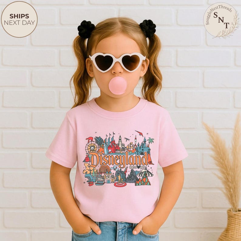 May include: A light pink t-shirt featuring a colorful Disneyland graphic. The graphic includes various park attractions and the Disneyland Resort text. The shirt is worn by a child wearing heart-shaped sunglasses and a bubblegum bubble.