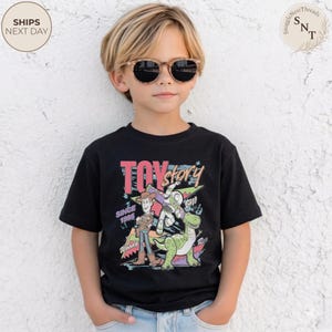 Może przedstawiać: Czarny t-shirt z kolorową grafiką postaci z Toy Story, w tym Chudego, Buzz'a Astrala i dinozaura. Wyświetlony jest r&oacute;wnież tekst "TOY STORY" i "SINCE 1995". Dziecko ma na sobie okulary przeciwsłoneczne.