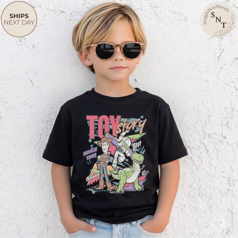 Może przedstawiać: Czarny t-shirt z kolorową grafiką postaci z Toy Story, w tym Chudego, Buzz'a Astrala i dinozaura. Wyświetlony jest r&oacute;wnież tekst "TOY STORY" i "SINCE 1995". Dziecko ma na sobie okulary przeciwsłoneczne.