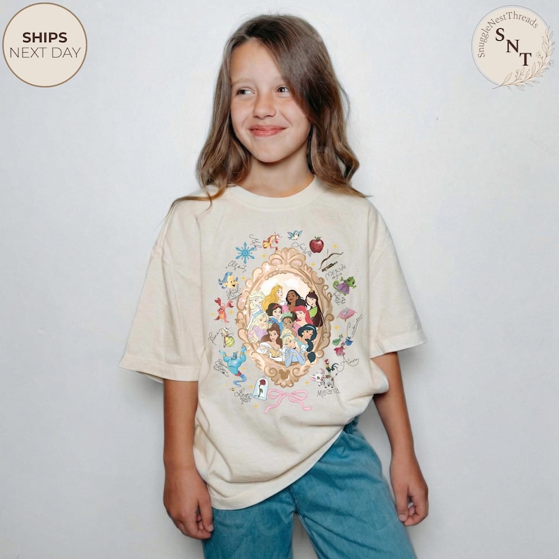 May include: Cream-coloured t-shirt featuring a gold-framed portrait of animated princesses. The shirt has a crew neck and short sleeves. The design includes various cartoon elements and the text "SnuggleNestThreads".