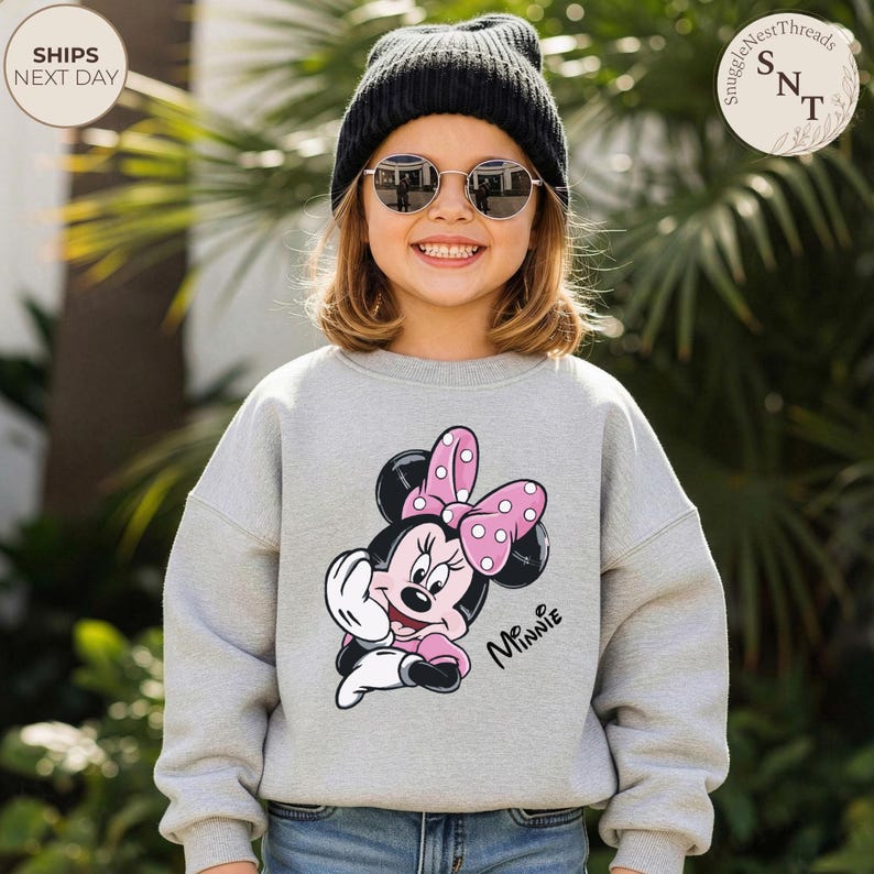 May include: A gray sweatshirt featuring a Minnie Mouse graphic with a pink bow and polka dots. The word "Minnie" is written in cursive. The person is wearing a black beanie and sunglasses.