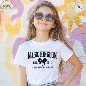 May include: A white t-shirt with the text "MAGIC KINGDOM" in black, with "EST. 1971" and "WALT DISNEY WORLD" below. The shirt is worn by a person wearing sunglasses. The background is a colorful mural.
