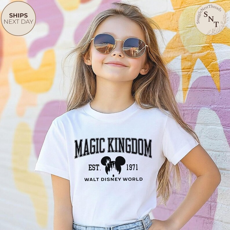 May include: A white t-shirt with the text "MAGIC KINGDOM" in black, with "EST. 1971" and "WALT DISNEY WORLD" below. The shirt is worn by a person wearing sunglasses. The background is a colorful mural.