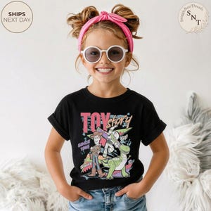 Może przedstawiać: Czarny t-shirt z kolorową grafiką postaci z filmu "Toy Story", w tym Woody, Buzz Lightyear i Rex. Projekt zawiera napis "Toy Story Since 1995". T-shirt jest noszony przez dziecko.