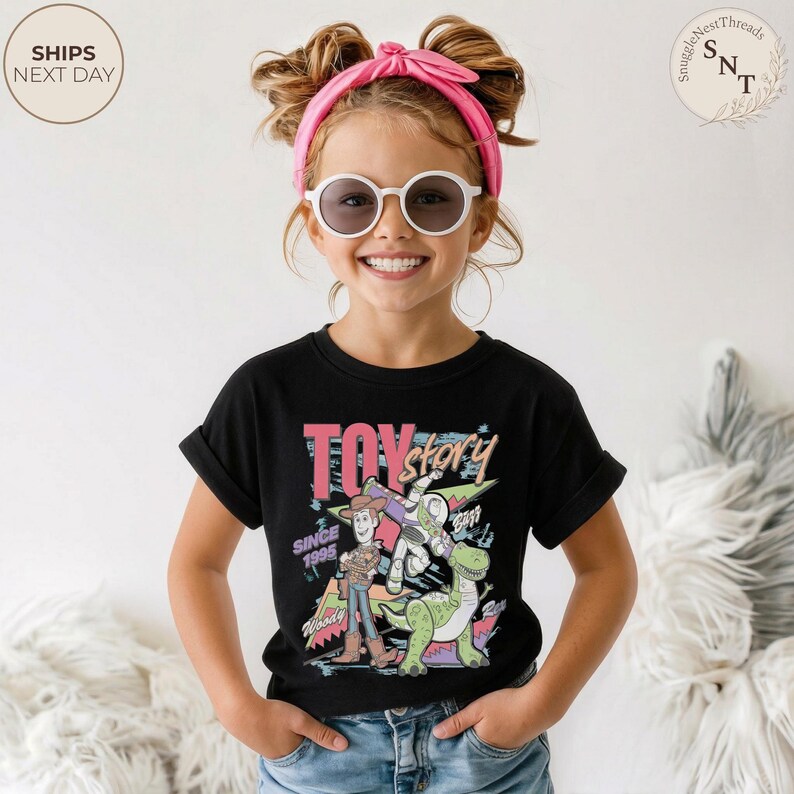 Może przedstawiać: Czarny t-shirt z kolorową grafiką postaci z filmu "Toy Story", w tym Woody, Buzz Lightyear i Rex. Projekt zawiera napis "Toy Story Since 1995". T-shirt jest noszony przez dziecko.