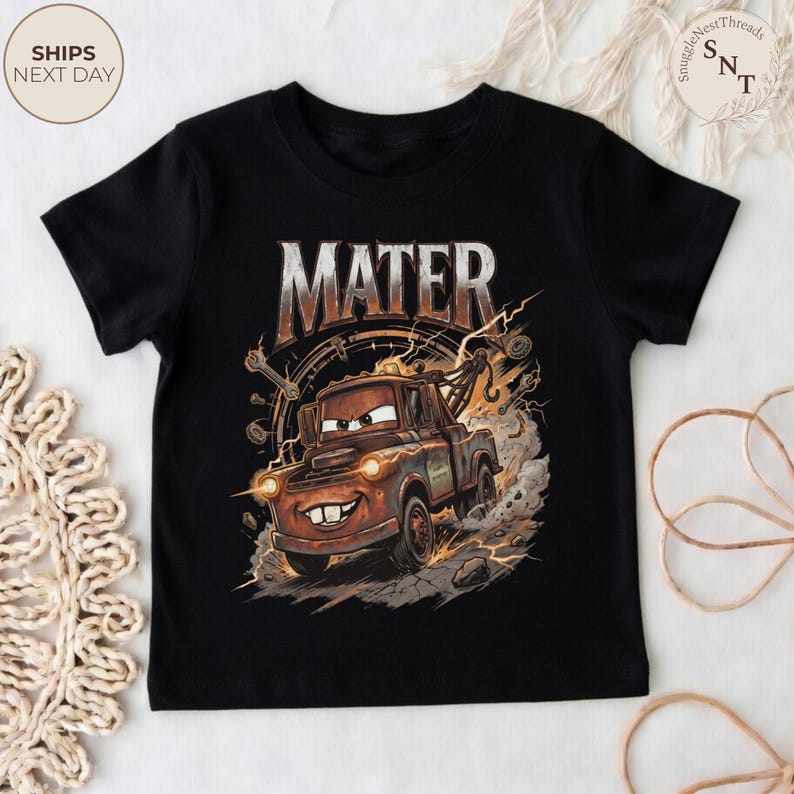 Puede incluir: Camiseta negra con una ilustraci&oacute;n de dibujos animados de Mater de la pel&iacute;cula Cars. El dise&ntilde;o incluye el nombre "MATER" en una fuente desgastada, con una imagen detallada de Mater, rodeado de chispas y elementos mec&aacute;nicos.