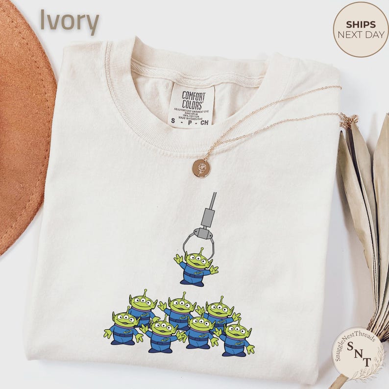 May include: Ivory-colored t-shirt featuring a graphic of green aliens in blue uniforms being lifted by a claw machine. The shirt has a crew neck and short sleeves. A gold necklace is draped across the shirt. The shirt is labeled "Comfort Colors".