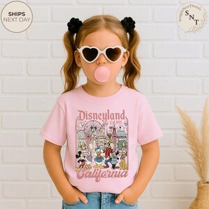 May include: A light pink short-sleeve t-shirt featuring a graphic print of Disneyland California with Mickey Mouse, Minnie Mouse, Donald Duck, Goofy, and Pluto. The design includes a Ferris wheel, castle, and the text Disneyland Est 1955 California.