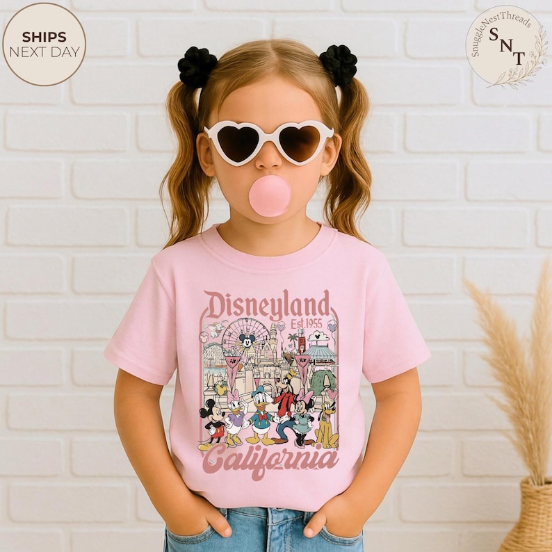 May include: A light pink short-sleeve t-shirt featuring a graphic print of Disneyland California with Mickey Mouse, Minnie Mouse, Donald Duck, Goofy, and Pluto. The design includes a Ferris wheel, castle, and the text Disneyland Est 1955 California.