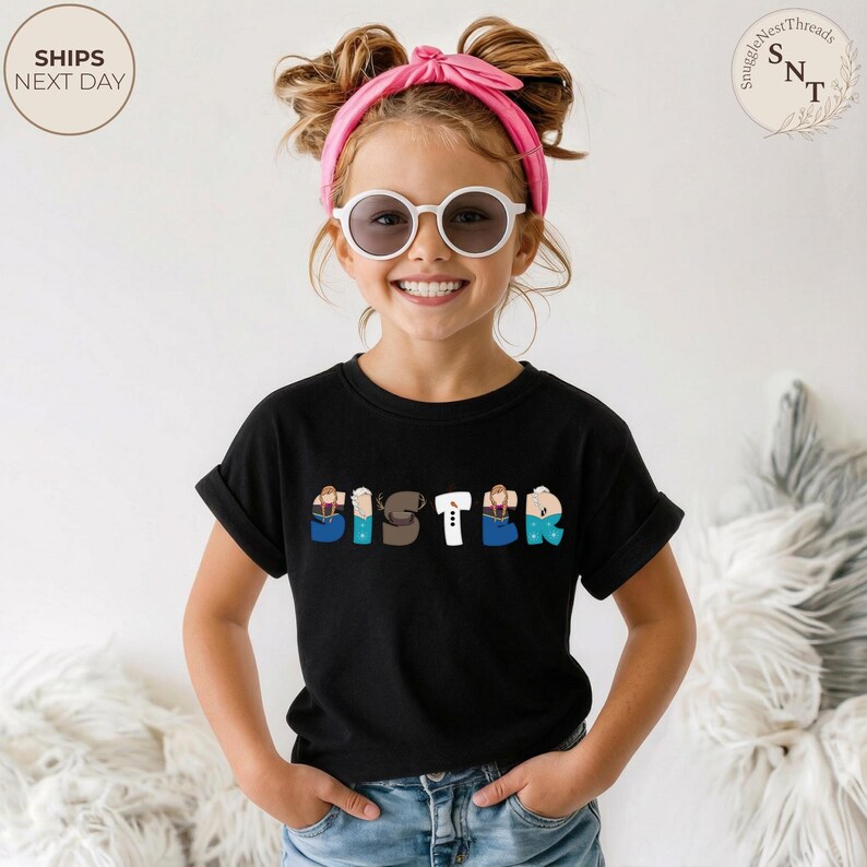 K&ouml;nnte beinhalten: Ein schwarzes T-Shirt mit dem Wort "SISTER" in bunten, cartoonartigen Buchstaben. Die Buchstaben sind so gestaltet, dass sie wie Personen aussehen. Das T-Shirt wird von einer jungen Person mit Sonnenbrille und einem rosa Stirnband getragen.