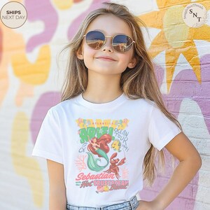 May include: A white short-sleeved t-shirt featuring a colorful graphic print of Ariel, Flounder, and Sebastian. The text reads Atlantica Ocean Tour 1991, Ariel, Sebastian with the Hot Crustacean Band. The design includes coral and musical notes.