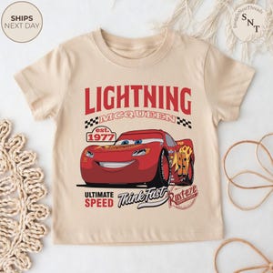 Op de afbeelding: Een beige T-shirt met korte mouwen met een rode cartoon Lightning McQueen raceauto-afbeelding. Het ontwerp bevat de tekst "LIGHTNING MCQUEEN" boven de auto en "ULTIMATE SPEED Think Fast" eronder. Het shirt heeft ook de tekst "est. 1977".