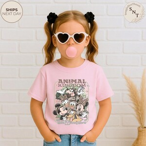 Pu&ograve; includere: Una maglietta rosa chiaro con una grafica di personaggi Disney, il testo "ANIMAL KINGDOM" e "Walt Disney World". La maglietta &egrave; indossata da una giovane persona con occhiali da sole a forma di cuore e ha una bolla di gomma da masticare.