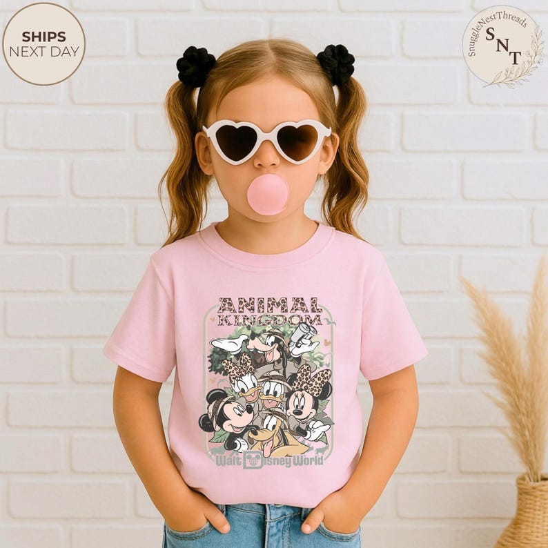 Pu&ograve; includere: Una maglietta rosa chiaro con una grafica di personaggi Disney, il testo "ANIMAL KINGDOM" e "Walt Disney World". La maglietta &egrave; indossata da una giovane persona con occhiali da sole a forma di cuore e ha una bolla di gomma da masticare.