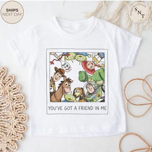 May include: White t-shirt featuring a colorful illustration of characters from the movie "Toy Story" within a framed design. The text "YOU'VE GOT A FRIEND IN ME" is printed below the image. A children's apparel item.