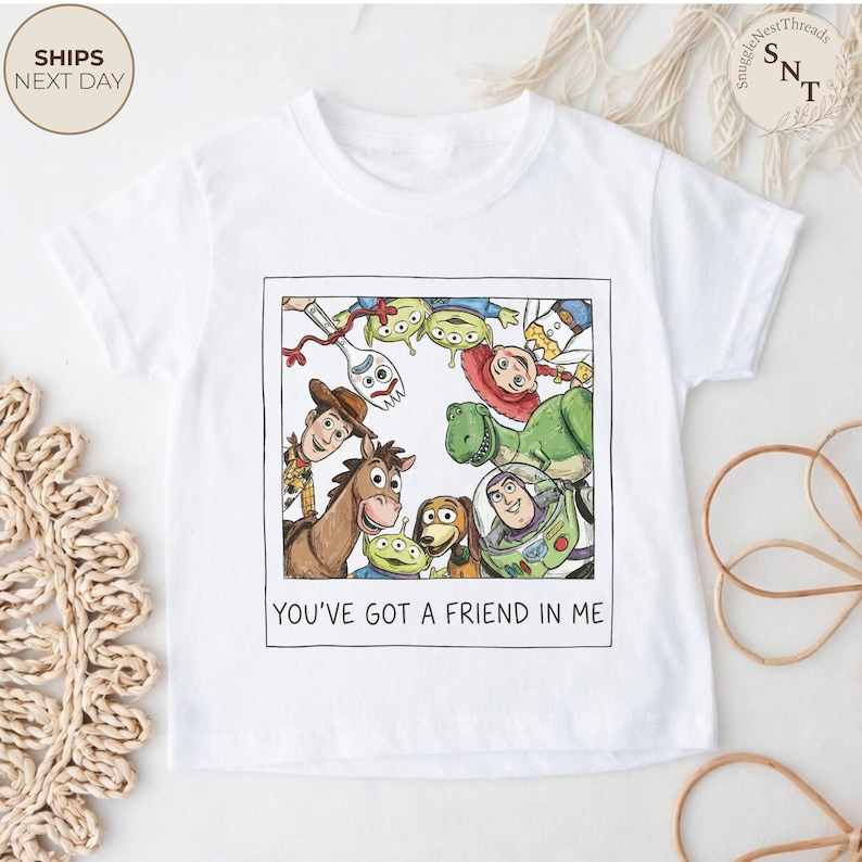 May include: White t-shirt featuring a colorful illustration of characters from the movie "Toy Story" within a framed design. The text "YOU'VE GOT A FRIEND IN ME" is printed below the image. A children's apparel item.