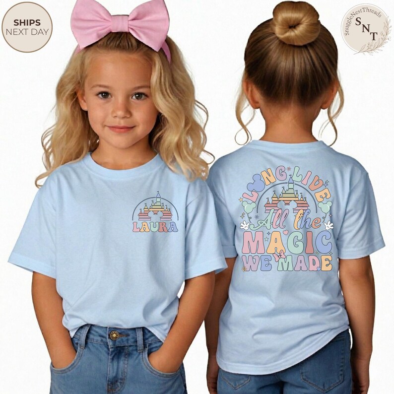 May include: Light blue t-shirts with a colorful castle graphic. One shirt has the name "LAURA" below the castle. The other shirt has the text "LONG LIVE All the MAGIC WE MADE" in pastel colors. Both shirts are short-sleeved.