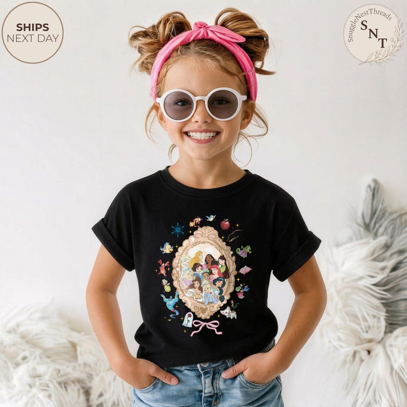 May include: A black t-shirt featuring a framed illustration of Disney princesses. The illustration is surrounded by decorative elements. The girl in the image is wearing white sunglasses and a pink headband. The shirt has rolled-up sleeves.