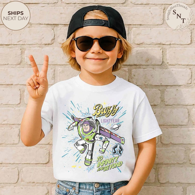 Puede incluir: Una camiseta blanca con un dise&ntilde;o de Buzz Lightyear, con el texto "Buzz Lightyear" y "To Infinity and Beyond". El dise&ntilde;o incluye detalles en verde, morado y azul. El ni&ntilde;o lleva una gorra negra y gafas de sol.