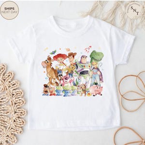 May include: White t-shirt featuring a colorful watercolor illustration of characters from the Toy Story movie franchise. The design includes Woody, Buzz Lightyear, Jessie, and other beloved characters. The shirt has a crew neck and short sleeves.