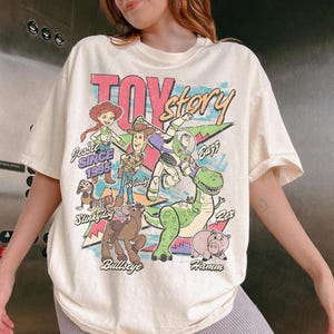 Puede incluir: Una camiseta de manga corta color crema con un estampado gr&aacute;fico colorido de los personajes de Toy Story, incluyendo a Jessie, Woody, Buzz Lightyear, Slinky Dog, Bullseye, Rex y Hamm. El texto dice Toy Story, Jessie Since 1995 y los nombres.