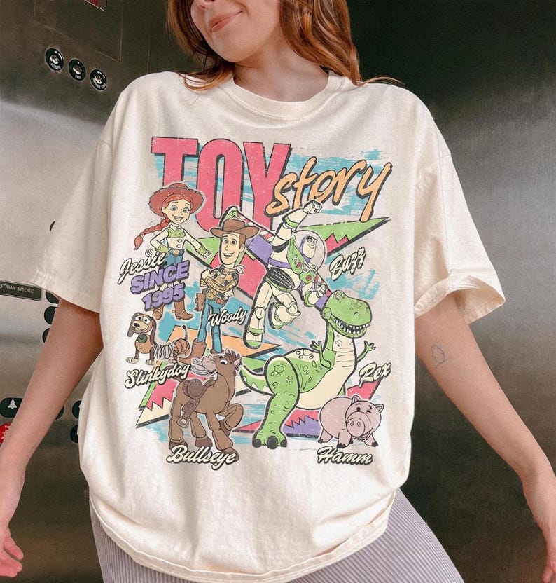 Puede incluir: Una camiseta de manga corta color crema con un estampado gr&aacute;fico colorido de los personajes de Toy Story, incluyendo a Jessie, Woody, Buzz Lightyear, Slinky Dog, Bullseye, Rex y Hamm. El texto dice Toy Story, Jessie Since 1995 y los nombres.