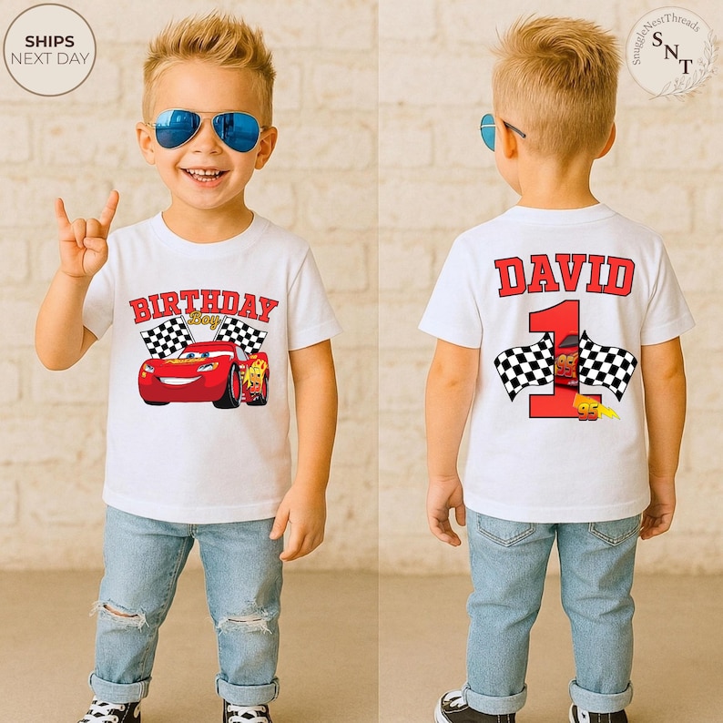 May include: A white t-shirt featuring the text "BIRTHDAY Boy" with a red race car graphic and checkered flags. The back of the shirt displays the name "DAVID" above a large red number "1" with more race car graphics and checkered flags. The child is wearing blue jeans.