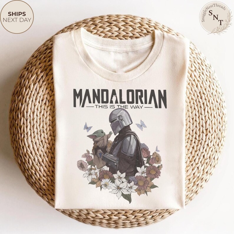 Puede incluir: Una camiseta de color crema con un gr&aacute;fico del Mandaloriano y Grogu rodeados de varias flores y mariposas. El texto "MANDALORIAN" y "THIS IS THE WAY" est&aacute; impreso en negro sobre la ilustraci&oacute;n central de los personajes.