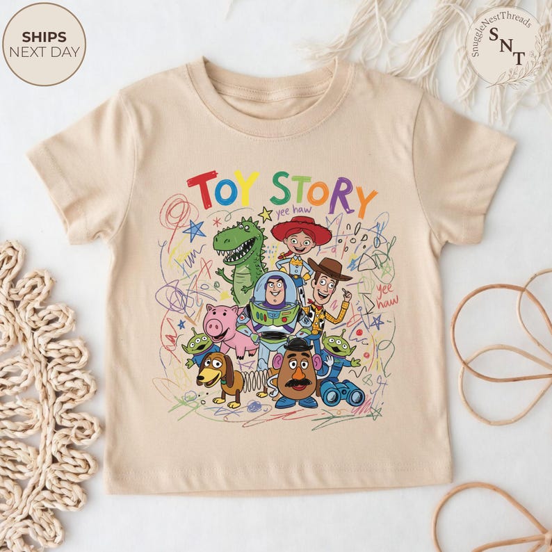 May include: A beige short-sleeved t-shirt featuring a colorful graphic of Toy Story characters and the words "TOY STORY" in a playful font. The design includes Woody, Buzz Lightyear, and other characters.