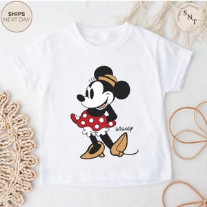 May include: White t-shirt featuring a cartoon illustration of Minnie Mouse in a red polka dot skirt and gold shoes. The Disney logo is printed on the shirt. The shirt has short sleeves.