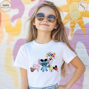 Pu&ograve; includere: T-shirt bianca con un personaggio dei cartoni animati che tiene in mano palloncini, zucchero filato e coni gelato. Il personaggio &egrave; blu con grandi occhi e orecchie nere. La maglietta &egrave; indossata da una persona con occhiali da sole.