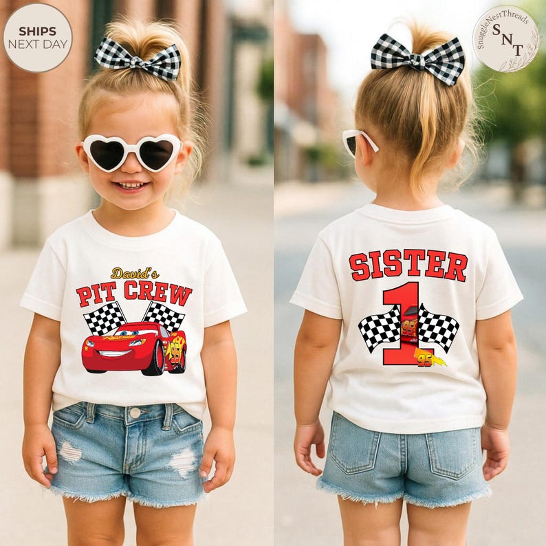 May include: White t-shirt with a red race car graphic and the text "David's PIT CREW". The back of the shirt says "SISTER 1" in red with checkered flags. The child is wearing denim shorts and heart-shaped sunglasses.