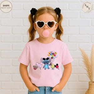 Pu&ograve; includere: Una maglietta rosa chiaro con un'illustrazione di Stitch, palloncini e coni gelato. Il design include zucchero filato e altri palloncini di personaggi Disney. La maglietta &egrave; indossata da un bambino con occhiali da sole a forma di cuore.