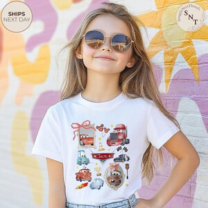 May include: A white t-shirt featuring colorful illustrations of cars, including a red race car, a blue car, and a truck. The word "Cars" is printed in the center. The shirt is worn by a young person wearing sunglasses.