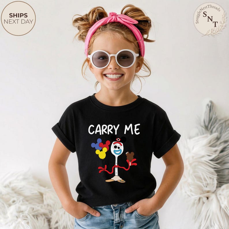 May include: Black t-shirt with the text "CARRY ME" and a cartoon character design. The design includes a fork-like character holding balloons and a chocolate treat. The child is wearing white sunglasses and a pink headband.